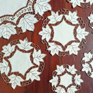 May include: A set of white and brown leaf-patterned doilies and a larger placemat. The doilies are circular with a white centre and a brown border featuring leaf designs. The placemat has a similar design. The items are arranged on a dark wooden surface.
