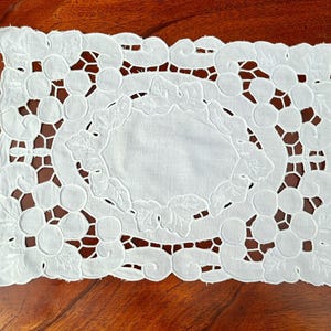Puede incluir: Tapete rectangular blanco con un diseño bordado de uvas y hojas. El centro presenta un óvalo con un patrón bordado similar. Los bordes son festoneados y el fondo es una superficie de madera oscura.
