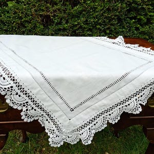 May include: A white rectangular tablecloth with a decorative crocheted lace border. The tablecloth has a stitched border and is displayed on a dark wooden table. The background features green foliage.