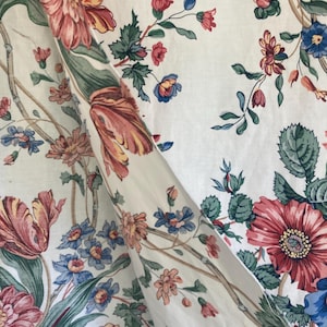 Puede incluir: Tela floral crema con un patrón repetido de grandes flores estilizadas. Las flores son en tonos de rojo, rosa, azul y amarillo, con hojas y tallos verdes. La tela está doblada, mostrando el patrón.