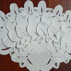 May include: A collection of white, intricately embroidered doilies. Each doily features a floral design with cut-out details and a scalloped edge. The doilies are arranged on a dark wooden surface, showcasing their delicate craftsmanship.