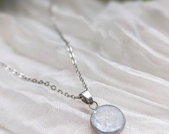 May include: A silver-toned necklace with a circular pendant. The pendant has a white, textured surface and is set in a silver bezel. The chain is delicate and rests on a white, textured fabric.