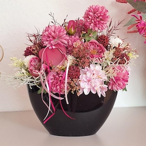 Könnte beinhalten: Ein Blumenarrangement mit verschiedenen rosa und magentafarbenen Blüten, darunter Chrysanthemen und Hortensien, in einer glänzenden, schalenförmigen schwarzen Vase. Ein kleines rosa Herzornament hängt mit dünnen Bändern an den Blumen.
