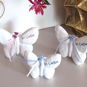 Könnte beinhalten: Drei weiße Keramik-Schmetterlingsfiguren mit dem Aufdruck "In Liebe". Jeder Schmetterling hat Bänder und eine kleine Blume. Die Schmetterlinge stehen auf einer hellen Oberfläche.