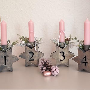 Könnte beinhalten: Vier sternförmige Kerzenhalter mit den Zahlen 1-4. Jeder Halter hat eine rosa Kerze, Grünzeug und dekorative Akzente. Die Halter sind aus Metall, ideal für die Weihnachtsdekoration.