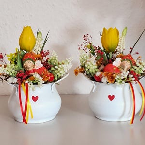 Könnte beinhalten: Zwei weiße Keramik-Blumentöpfe, jeweils mit einem roten Herz verziert und mit bunten Blumen gefüllt, darunter gelbe Tulpen. Jeder Topf hat eine kleine Figur und dekorative Bänder.