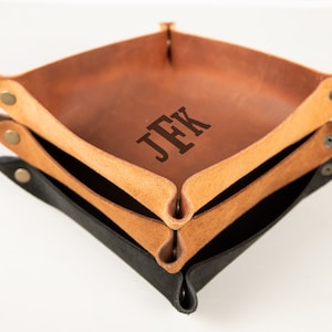 May include: Three leather valet trays in shades of brown and black are stacked. The top tray is a medium brown with the initials "JFK" engraved. The middle tray is a lighter tan color, and the bottom tray is black. Each tray has snap closures.