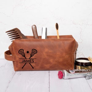 May include: Brown leather toiletry bag with a zipper closure. The bag has a personalized design with two lacrosse sticks crossed over each other and the name "JAMES" printed below.