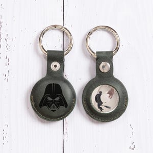 May include: Two dark green leather keychains with silver rings. One keychain features a black and white image of Darth Vader's face. The other keychain features a silver disc with a black and white image of a person.
