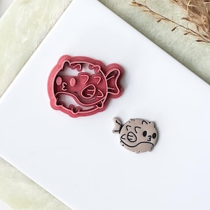 May include: A pink plastic pufferfish-shaped cookie cutter alongside a small, light brown pufferfish-shaped cutout. The cookie cutter features a detailed outline of the fish, while the cutout displays a smooth surface with a printed fish face.