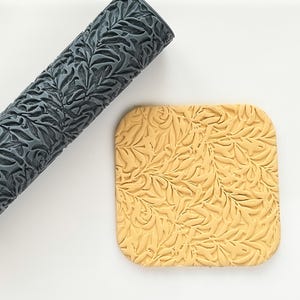 May include: A grey patterned rolling pin and a yellow square tile with a leaf design. The rolling pin is cylindrical and features a repeating leaf pattern. The tile has rounded corners and a similar leaf pattern.