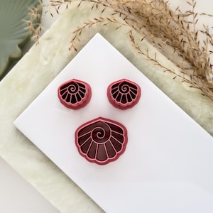 May include: Three seashell-shaped polymer clay cutters in varying sizes. The cutters are a deep red color with a spiral design in the center. They are arranged on a white surface with a green and beige background.