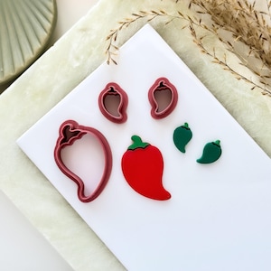 May include: A collection of red and green chili pepper-shaped cookie cutters and a red chili pepper. The cookie cutters are in the shape of a chili pepper. The chili pepper is red with a green stem. The items are on a white surface.
