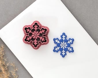May include: A pink snowflake-shaped cookie cutter with a detailed floral center and a blue snowflake-shaped cookie with white embossed details. The cookie cutter features a raised edge and a hollow interior, while the cookie displays a flat surface.