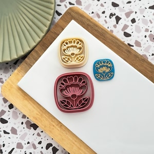 May include: Three floral-shaped cookie cutters in varying sizes and colours. One is beige, one is burgundy, and the smallest is blue with gold accents. The cutters are on a white surface with a wooden and patterned background.