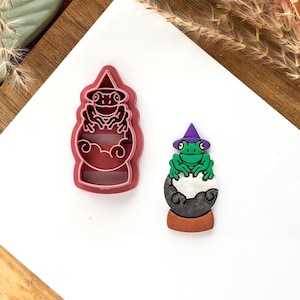 May include: A red plastic cookie cutter and a painted frog figurine. The cookie cutter is shaped like a frog in a crystal ball wearing a witch hat. The frog figurine is green with a purple hat and sits on a gray ball.