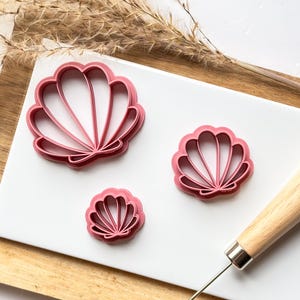 May include: Three pink plastic seashell cookie cutters of different sizes on a white surface. The largest cutter has a detailed design with multiple ridges. The cutters are next to a wooden rolling pin.