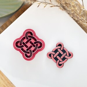 May include: Two pink and black Celtic knot-shaped cookie cutters. One is larger with a pink border and a dark pink interior. The other is smaller with a pink border and a black interior. Both are on a white surface.