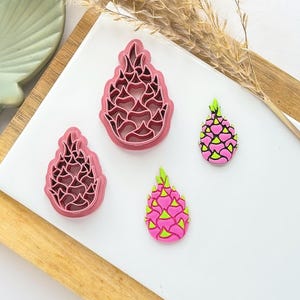 May include: Two pink dragon fruit cookie cutters and two dragon fruit-shaped decorations. The cookie cutters are in the shape of a dragon fruit, with a detailed interior design. The decorations are pink with green and yellow accents.