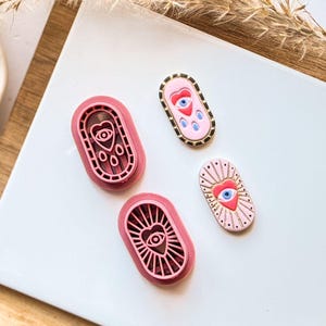 May include: Four pink and gold oval-shaped charms and stamps. Two pink stamps feature an eye and heart design. Two charms have an eye and heart design, one with gold trim and the other with sun rays.