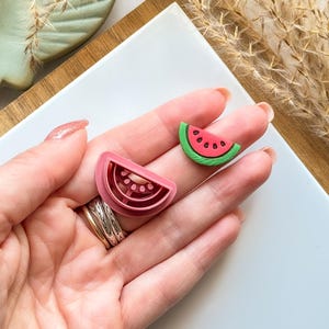 May include: A pink watermelon-shaped cookie cutter and a watermelon-shaped charm. The cookie cutter is made of pink plastic and has a detailed design. The charm is green, red, and black. The items are on a white surface.