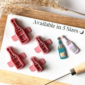 May include: Set of five champagne bottle cookie cutters in various sizes. The cutters are pink and feature a champagne bottle design. The text "Available in 5 Sizes" is written on the white surface.