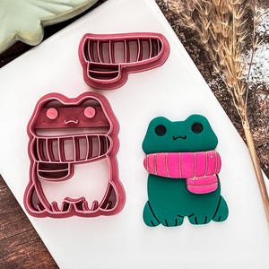 May include: Three frog-shaped cookie cutters. Two are pink plastic outlines, and one is a finished green frog with a pink scarf. The frog cutters are for baking and crafting.