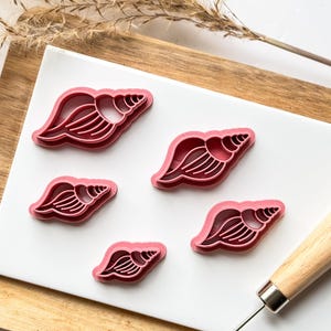 May include: Five pink plastic cookie cutters shaped like seashells on a white surface. The cutters are arranged in a staggered pattern. A wooden rolling pin is in the background.