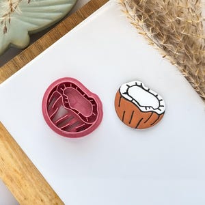 May include: A pink, coconut-shaped cookie cutter and a hand-painted coconut decoration. The cutter is plastic with a detailed interior. The coconut decoration is brown with a white interior and black outlines. The items are on a white surface.
