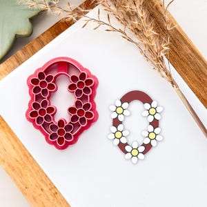 May include: Two cookie cutters in the shape of the letter "O" decorated with daisy flowers. The cutter on the left is red and the cutter on the right is brown with white daisy flowers.
