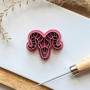 May include: A pink plastic cookie cutter shaped like a ram skull with detailed horns.