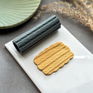 May include: A dark grey cylindrical rolling pin with a textured herringbone pattern and a small, rectangular, mustard yellow clay piece featuring the same embossed herringbone design, both resting on a smooth white rectangular surface.