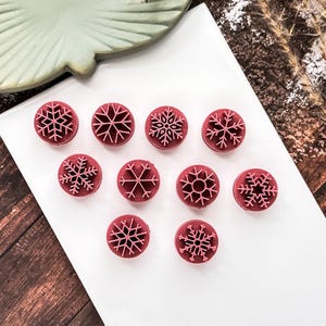 May include: Ten red, circular snowflake stamps are displayed on a white rectangular plate. Each stamp has a different snowflake design. The plate rests on a wooden surface, with a green decorative plate in the background.