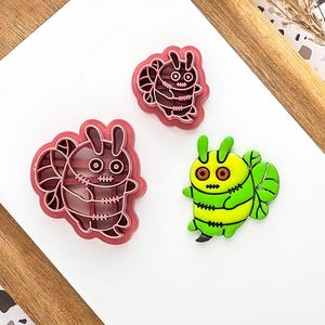 May include: Three novelty bug-shaped cookie cutters. Two are red with white detailing, and one is green and yellow. The cutters are on a white surface with a wooden border.