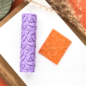 May include: A purple rolling pin with a ghost pattern and an orange square with a paisley design sit on a white surface. The rolling pin is cylindrical and covered in smiling ghost faces. The square has a raised paisley pattern.