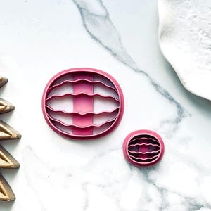 May include: Two pink plastic cookie cutters shaped like a wavy oval. The larger cutter has a wavy line design across the centre. The smaller cutter is a smaller version of the larger cutter.