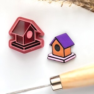 May include: A pink plastic cookie cutter shaped like a birdhouse, and a small orange and purple birdhouse with a pink base.