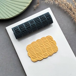 May include: A dark teal cylindrical rolling tool with a grid-like texture and a mustard yellow rectangular piece with rounded edges featuring a matching embossed grid pattern. The tool and the yellow piece are positioned on a white rectangular surface.