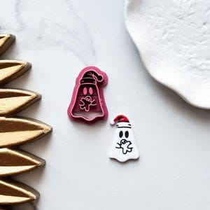 May include: A pink plastic cookie cutter shaped like a ghost wearing a Santa hat, beside a white ghost-shaped decoration with a Santa hat. The ghost has black eyes and a gingerbread man design. The cutter is on a white surface.