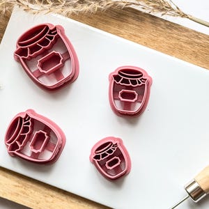 May include: Four pink plastic cookie cutters shaped like jars with lids and labels. The cutters are on a white surface with a wooden background.