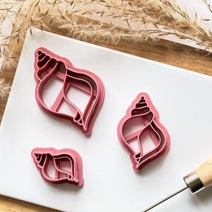 May include: Three pink plastic cookie cutters shaped like seashells. The cutters are on a white surface.