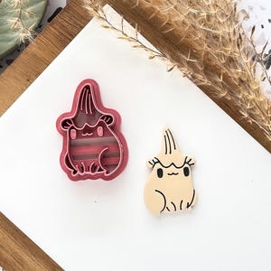 May include: A pink plastic cookie cutter and a beige cookie in the shape of a cartoon character with a tall, pointed head and a smiling face. The cookie cutter has a matching design with horizontal lines. The items are on a white surface.