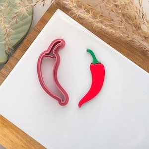 May include: A red chili pepper cookie cutter and a red chili pepper with a green stem rest on a white surface. The cookie cutter is a shade of pink. The items are on a white surface with a wooden and green background.