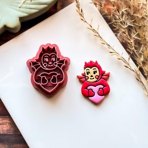 May include: Two devil-themed cookie cutters and a decorated cookie. The cookie cutters are red and shaped like a devil holding a heart. The decorated cookie is red with a pink heart.