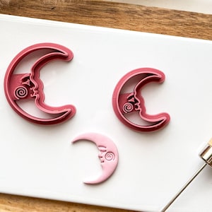 May include: Three pink crescent moon cookie cutters with a face design. The largest cutter is on the left, the smallest is on the right, and the middle cutter is in the centre.