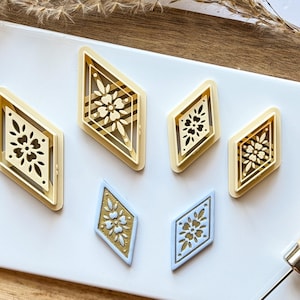 May include: Five diamond-shaped cookie cutters with intricate floral designs. The cutters are made of a light yellow plastic and are arranged on a white surface. Two of the cutters are a lighter blue colour.