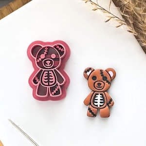 May include: A pink and brown teddy bear cookie cutter and a matching cookie. The bear has stitched details and a skeleton design on its body. The cookie cutter is approximately 3 inches tall.