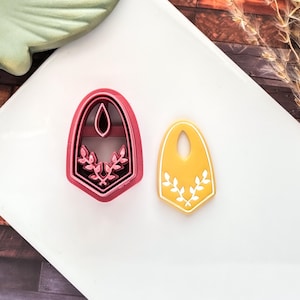 May include: Two cookie cutters. One is a dark pink, shield-shaped cutter with a leaf design. The other is a yellow, shield-shaped cookie with a white leaf design. Both have a teardrop-shaped hole at the top.