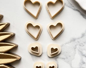 May include: A collection of beige heart-shaped cookie cutters in various sizes. The cutters are arranged on a white and gray marble surface, with a gold decorative element in the corner. Ideal for baking and crafting.