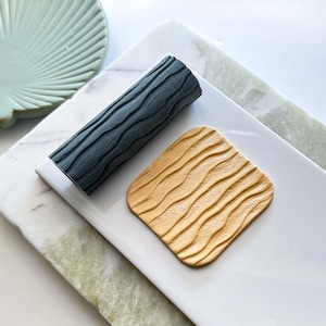 May include: A dark grey cylindrical roller with a wavy, textured surface alongside a square, light tan piece of material featuring the same embossed wavy pattern. Both items are positioned on a smooth, white rectangular surface.